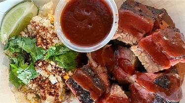 Aerial view of burnt ends with cheesy corn, a container of BBQ sauce and a lime wedge