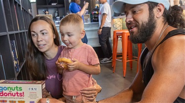 Family of three showing young boy holding a donut while picking out a donut-themed board game from the wall of games