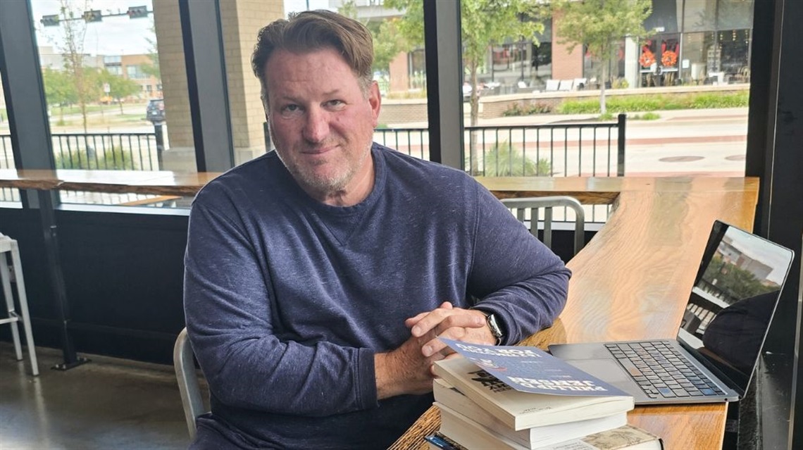 Brian W. sitting at wooden bar height table inside the Public Market with a stack of books and a laptop